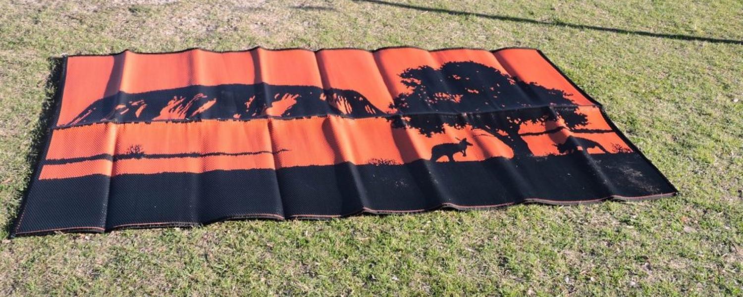 A Jawa Off Road Camper Trailer camping mat unfolded onto grass. "Outback" is a black and orange mat with a graphic of Uluru, a tree and kangaroos. Available in sizes suited to your caravan in Sunshine Coast Queensland.