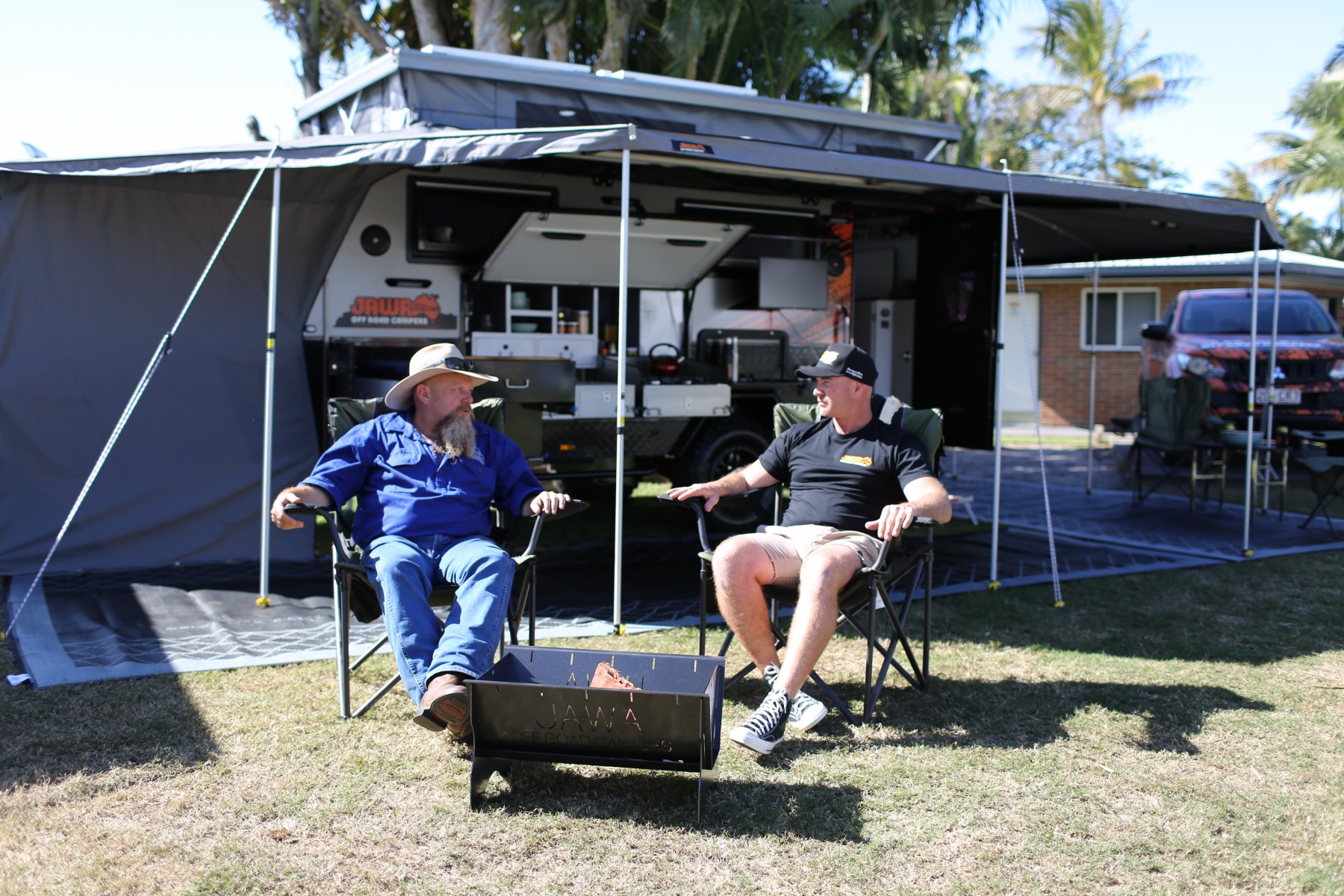 Jawa Sirocco fully set up at campsite with alfresco kitchen and external storage on hybrid caravan. Two men are sitting in front of the caravan in a park with a Jawa Off Road Camper Trailers fire pit. All are on sale in Sunshine Coast Queensland.
