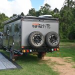 Jawa Infinity 15 hybrid off road camper, large camper for family of four, pictured towed by four wheel drive back view with camping mat next to it. Two spare tires on the back of camper.