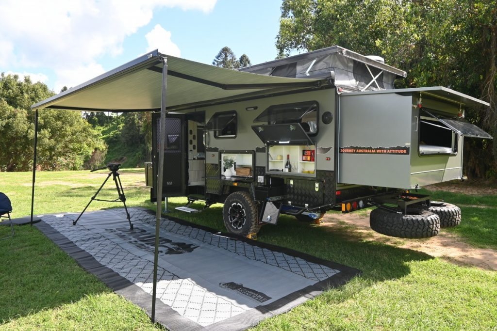Jawa Infinity 15 hybrid off road camper, large camper for family of four. Camper is pictured parked at grassy camping site with roof popped up and back extended, awning set up and camping mat laid out on entrance side.