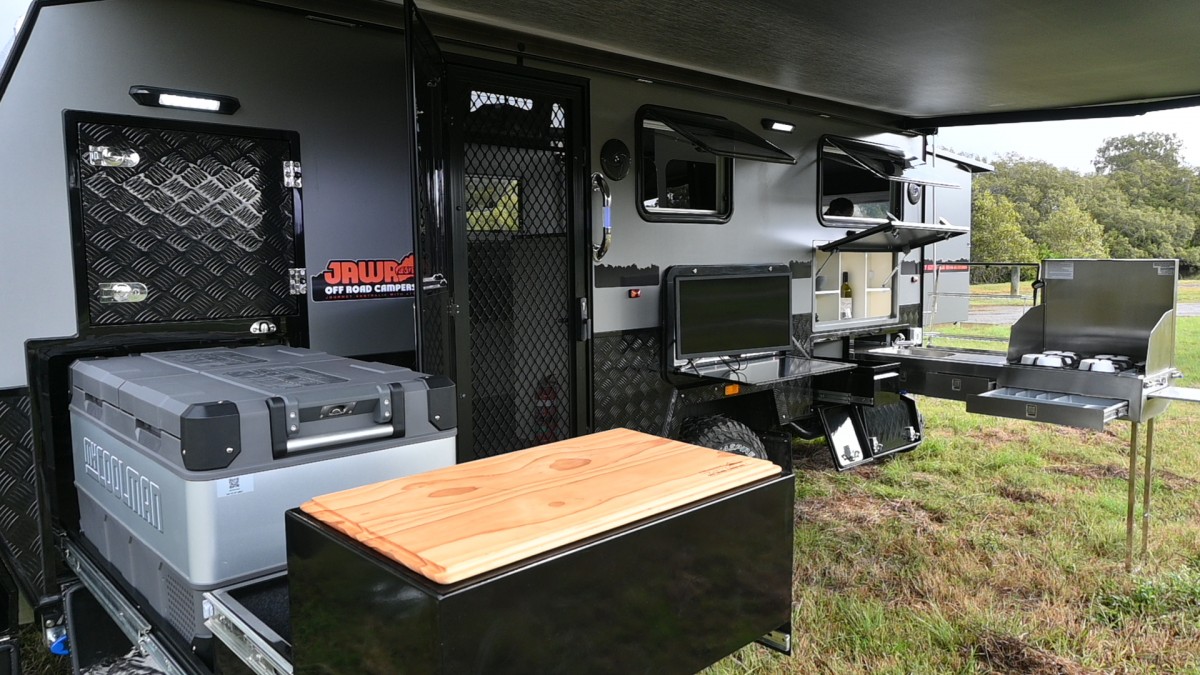 Jawa Infinity Escape hybrid off road camper trailer, large and spacious camper for couples, pictured under awning showing external fridge and storage extended out with chopping board, shelf with TV, external pantry and external kitchen with four burner cooktop, sink, bench and cutlery and cooking utensil storage drawer.