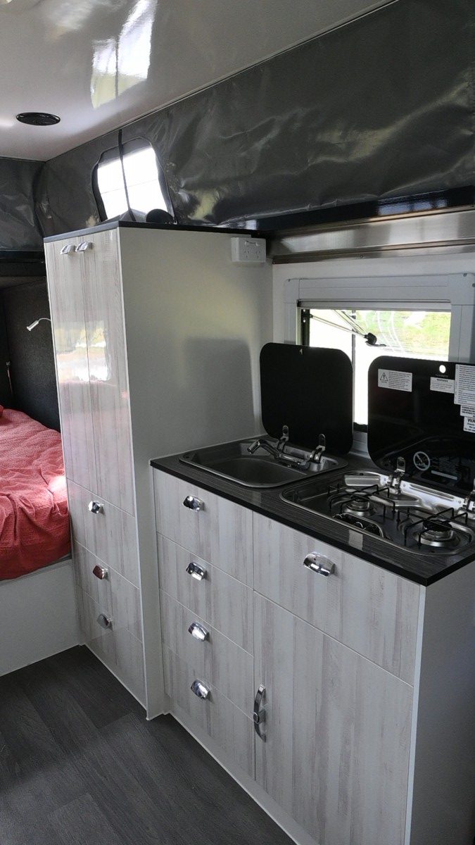 Jawa Infinity Escape hybrid off road camper trailer, large and spacious camper for couples, pictured inside camper showing wardrobe and cupboards with sink and two cooktop stove uncovered for use.