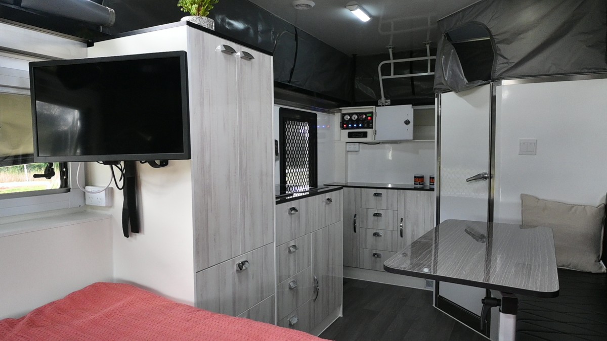 Jawa Infinity Escape hybrid off road camper trailer, large and spacious camper for couples, pictured inside camper with swing arm table in front of single sofa, TV mounted to wardrobe, cupboards with sink and cooktop, more storage on back wall and internal ensuite.