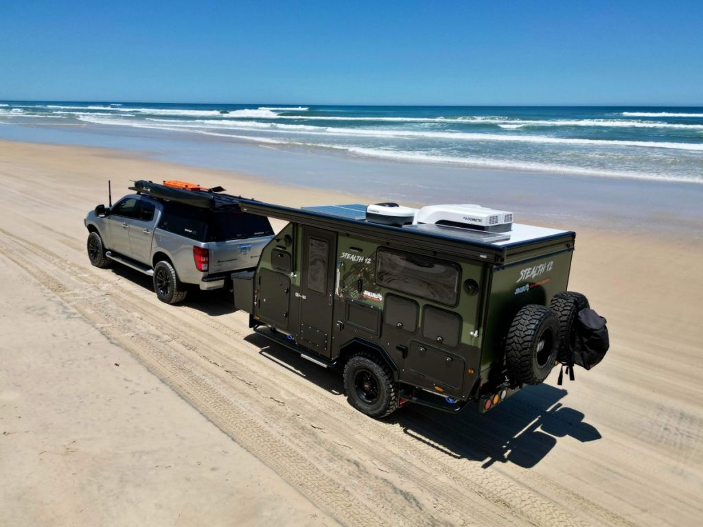 Jawa Stealth 12 tough hybrid off road camper packed with features, pictured towed across the beach by a four wheel drive.