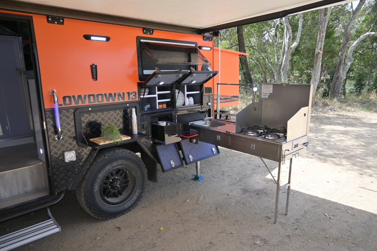 Jawa Lowdown 13 Cupboard low hybrid poptop off road camper with lots of storage perfect for adventuring couples and storing in standard garage, pictured under awning with three pantry compartments open, shelf and external kitchen with sink and four burner stove.