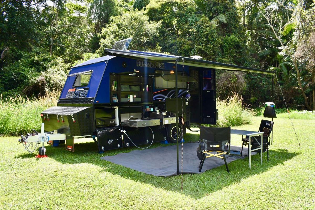 Jawa Commander hardtop off road camper with full setup. Camper trailer, awning and mat with storage drawers pulled out, pictured in a camping reserve with bushes and trees in the background.