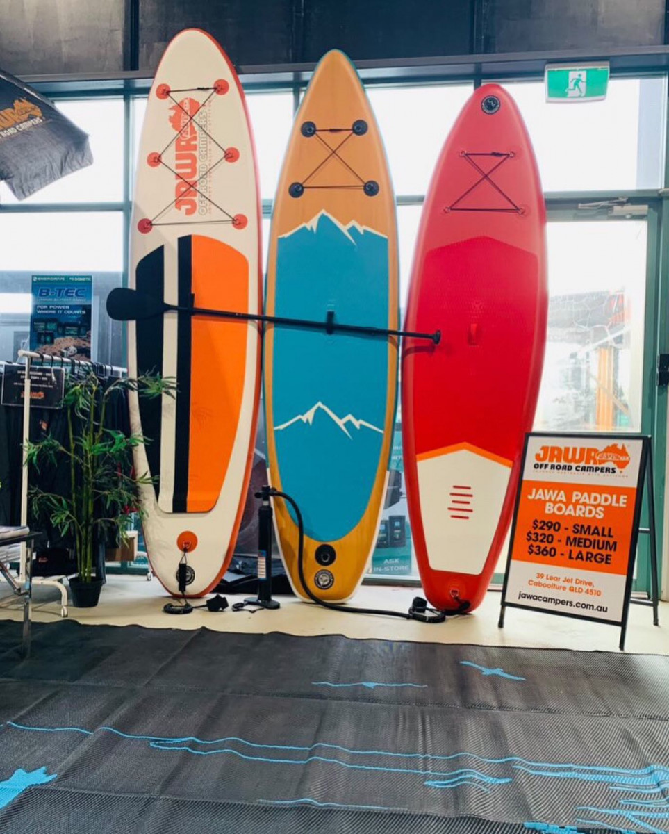 Three paddle boards from longest to shortest with a paddle attached horizontally across them. Large one is $360 orange, white and black. Medium is $320 and orange and blue. Small is $290 and pink and white. For sale at Jawa Off Road Camper Trailers in the Sunshine Coast Queensland.
