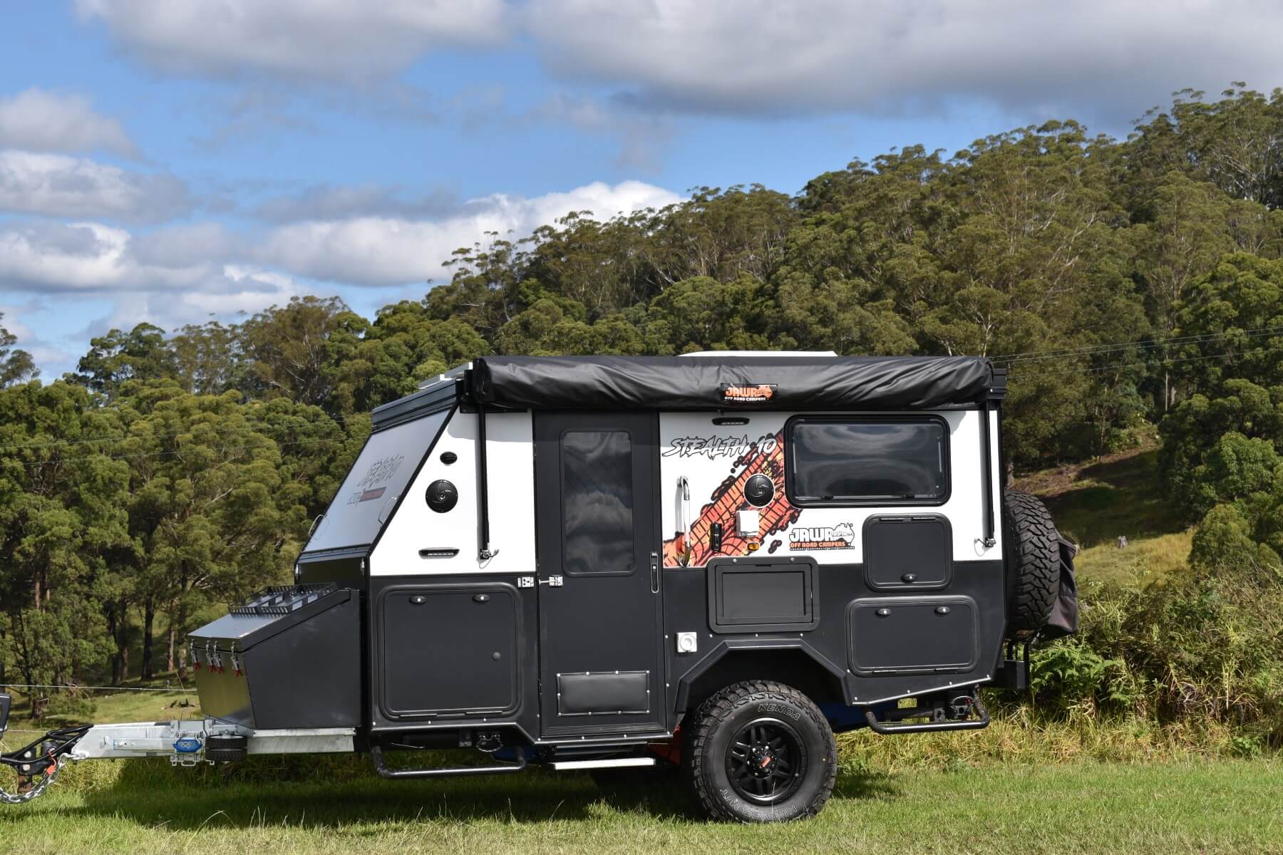 Jawa Stealth 10 hybrid compact off road camper trailer, small size camper for couples, parked on grass with hilly and bush landscape behind.